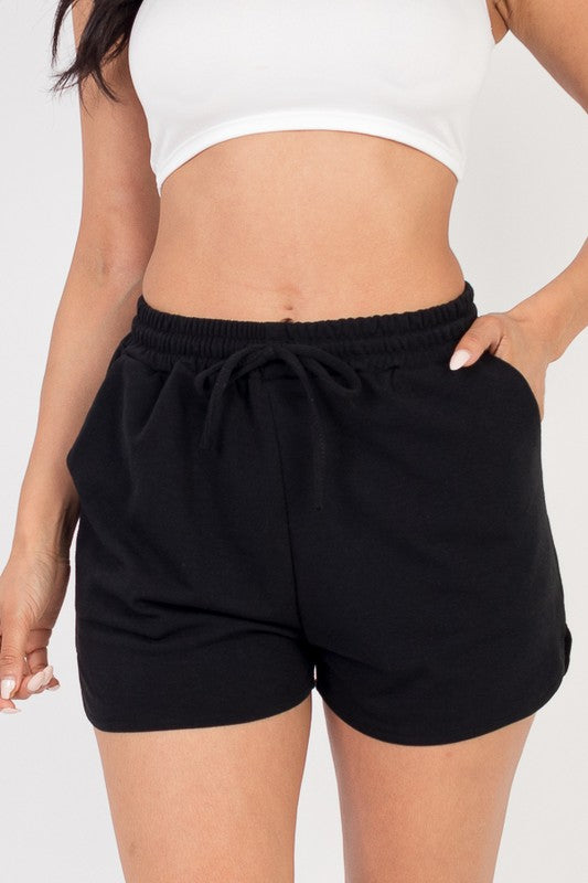 The image shows a woman wearing a white crop top and a black shorts. She's posing with one hand in her pocket and the other on her thigh, against a plain white background.