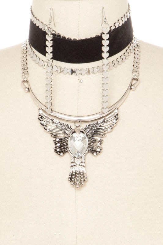 A stylish layered necklace on a mannequin. There's a black velvet choker with gold accents at the top, followed by multiple gold chain layers. The main piece is a gold necklace with a pendant featuring a clear gemstone surrounded by wing-like details and a tassel at the bottom.