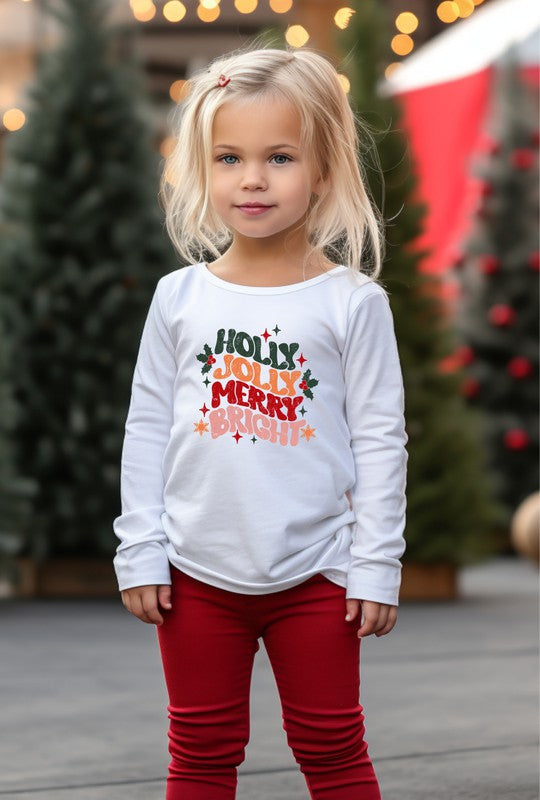 Young girl wearing a white long-sleeve shirt with Christmas-themed text, standing in front of decorated trees.