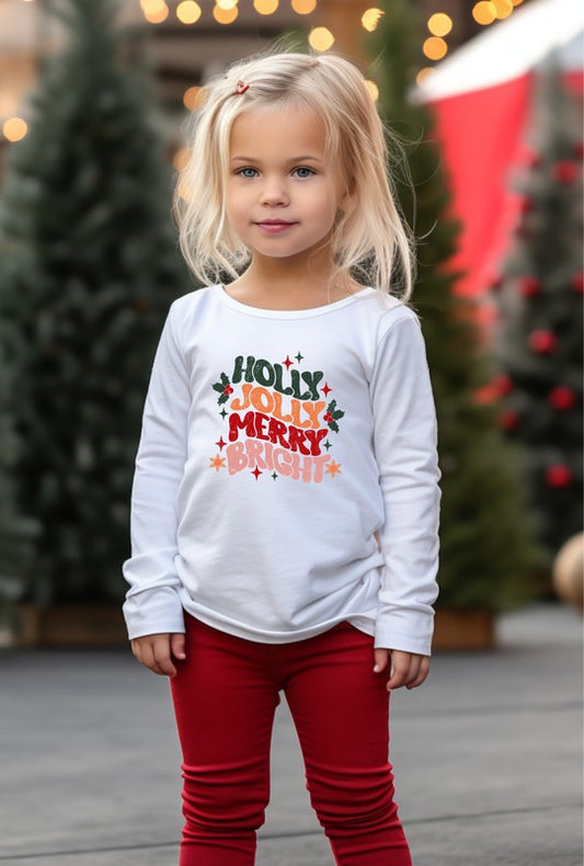 Young girl wearing a white long-sleeve shirt with Christmas-themed text, standing in front of decorated trees.