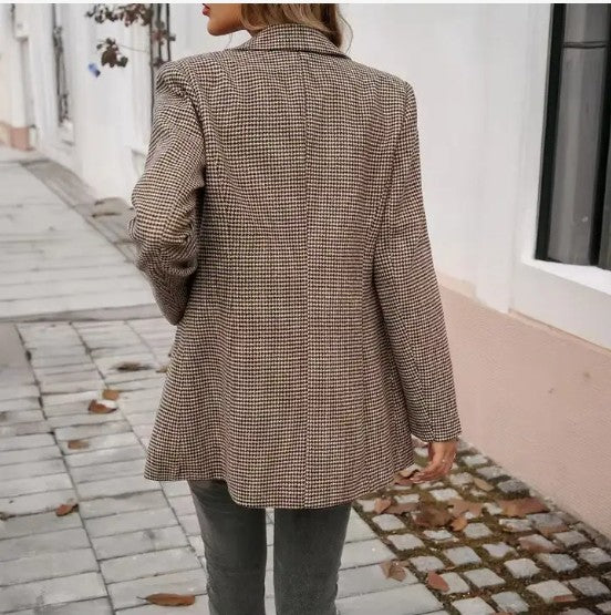 A lady in a brown Houndstooth blazer with a white top, grey jeans, and some dope accessories like a beige purse and gold jewelry. She's posing on a stylish cobblestone sidewalk with a white wall backdrop.