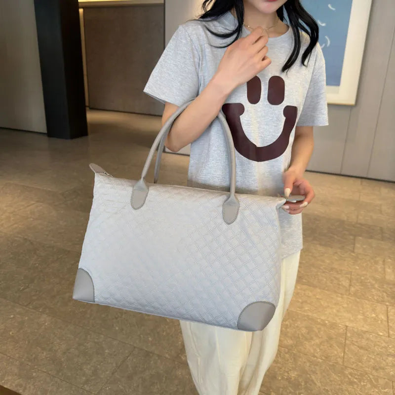 A person is standing indoors wearing a light gray t-shirt with a maroon smiley face and cream-colored pants, holding a large light gray quilted handbag with darker gray corner accents. The setting includes a tiled floor and a framed picture on the wall, creating a casual, stylish vibe.