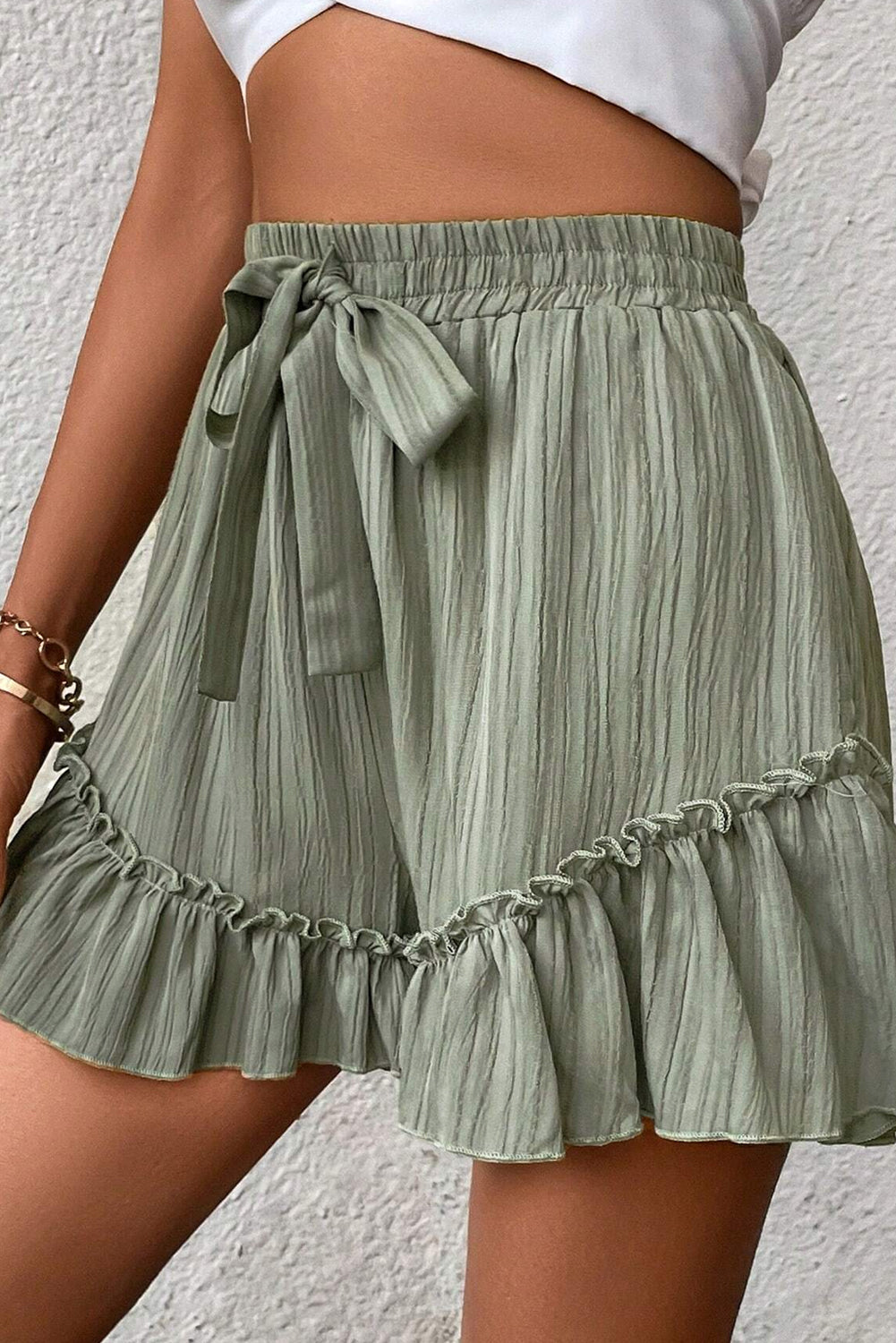 A woman wearing high-waisted, light green shorts with a drawstring waist and ruffled hem. She is dressed casually with a crop top, and her hand is resting on her hip, standing against a textured background.