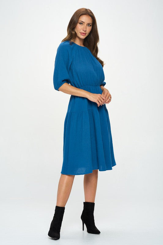 Woman wearing a cotton tiered midi dress with ¾ sleeves and a gathered waist, styled with black crisscross heels against a white background.