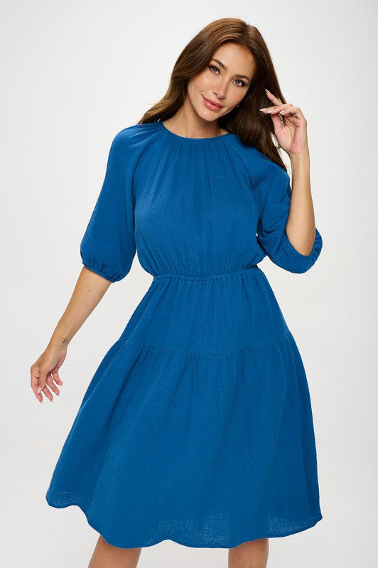 Woman wearing a cotton tiered midi dress with ¾ sleeves and a gathered waist, styled with black crisscross heels against a white background.