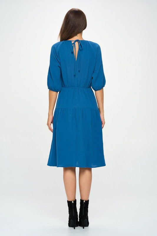 Woman wearing a cotton tiered midi dress with ¾ sleeves and a gathered waist, styled with black crisscross heels against a white background.