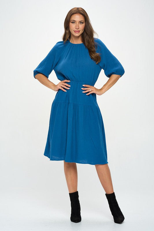 Woman wearing a cotton tiered midi dress with ¾ sleeves and a gathered waist, styled with black crisscross heels against a white background.