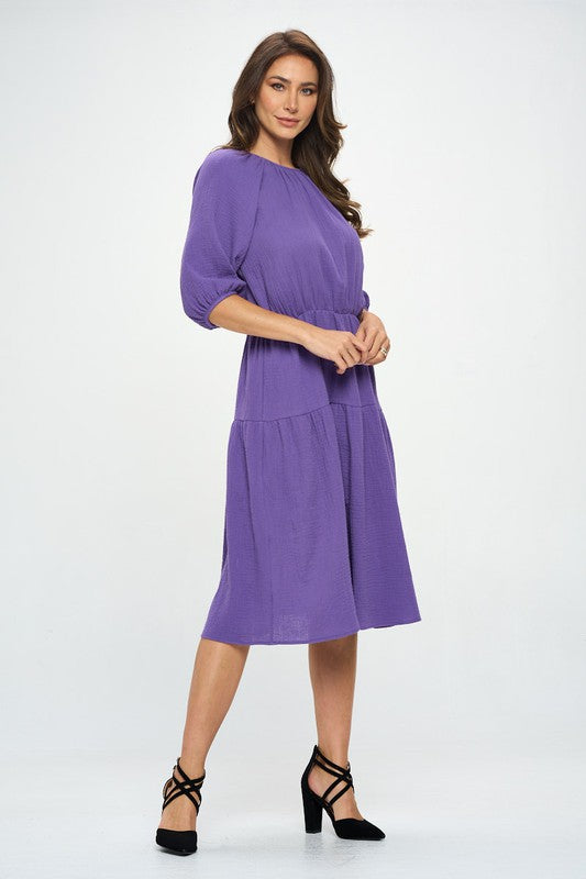 Woman wearing a purple tiered midi dress with ¾ sleeves and a gathered waist, styled with black crisscross heels against a white background.