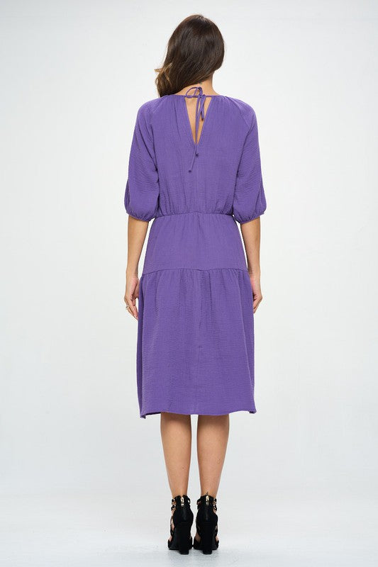 Woman wearing a purple tiered midi dress with ¾ sleeves and a gathered waist, styled with black crisscross heels against a white background.