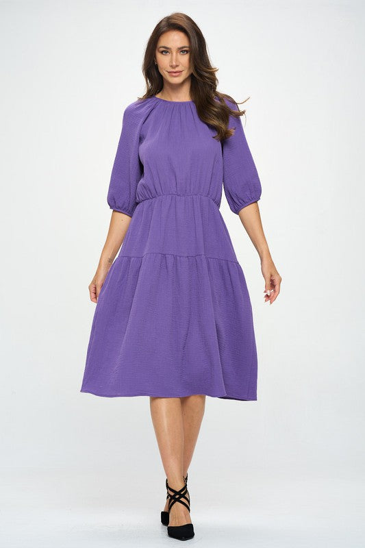 Woman wearing a purple tiered midi dress with ¾ sleeves and a gathered waist, styled with black crisscross heels against a white background.