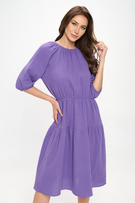 Woman wearing a purple tiered midi dress with ¾ sleeves and a gathered waist, styled with black crisscross heels against a white background.