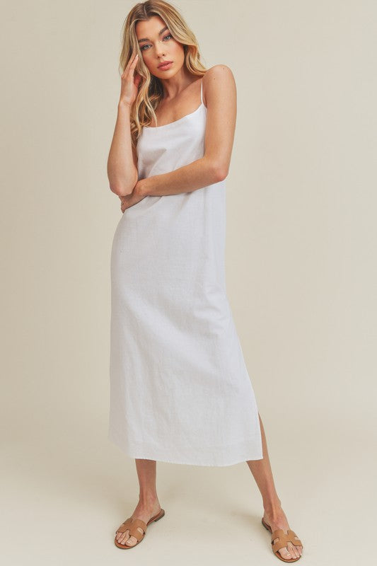Woman in a vibrant white dress striking a pose against a neutral beige background. The dress features thin straps and a side slit, complemented by her long blonde hair and silver jewelry.