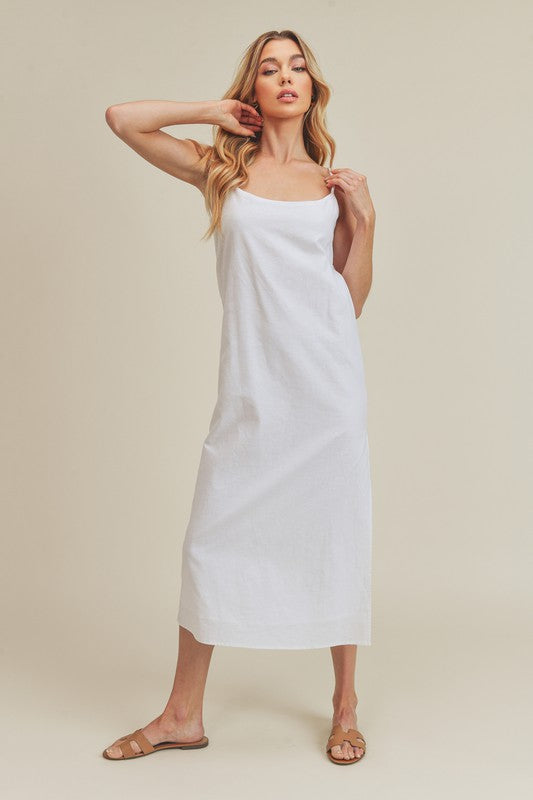 Woman in a vibrant white dress striking a pose against a neutral beige background. The dress features thin straps and a side slit, complemented by her long blonde hair and silver jewelry.