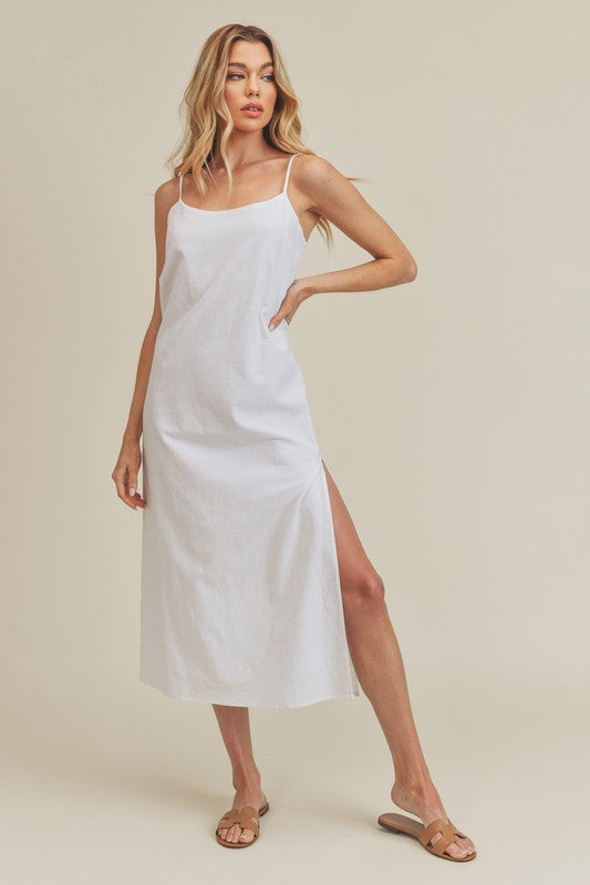 Woman in a vibrant white dress striking a pose against a neutral beige background. The dress features thin straps and a side slit, complemented by her long blonde hair and silver jewelry.