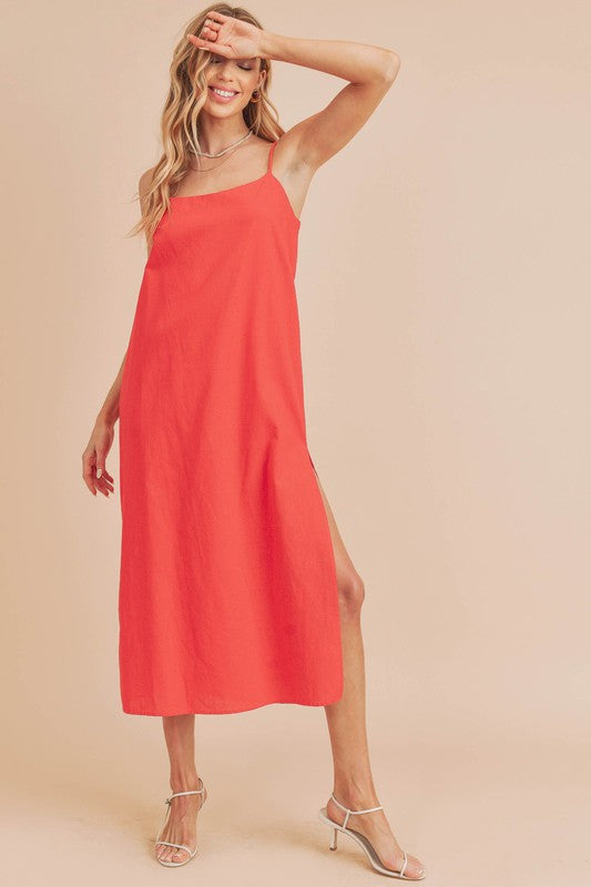 Woman in a vibrant red dress striking a pose against a neutral beige background. The dress features thin straps and a side slit, complemented by her long blonde hair and silver jewelry.