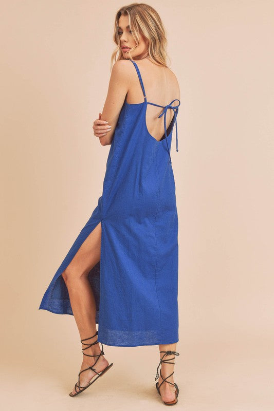 Woman in a vibrant blue dress striking a pose against a neutral beige background. The dress features thin straps and a side slit, complemented by her long blonde hair and silver jewelry.