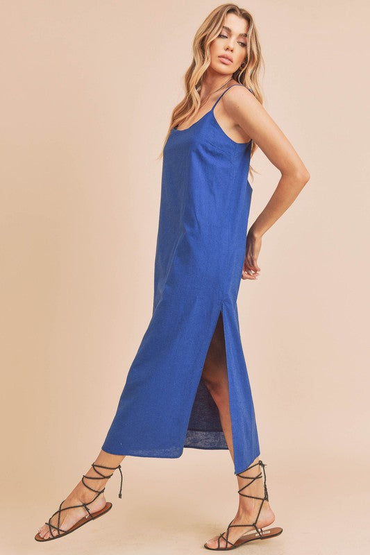 Woman in a vibrant blue dress striking a pose against a neutral beige background. The dress features thin straps and a side slit, complemented by her long blonde hair and silver jewelry.