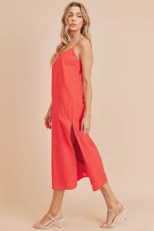 Woman in a vibrant red dress striking a pose against a neutral beige background. The dress features thin straps and a side slit, complemented by her long blonde hair and silver jewelry.