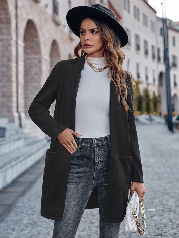 A woman posing on a cobblestone street. She's wearing a black hat, a long black cardigan over a white top, jeans, and has a white purse with a gold chain handle. Buildings are blurred in the background.