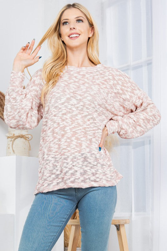 A woman posing in a casual outfit. She's wearing a gray and white patterned sweater and blue jeans. Her left hand is on her head, and her right hand is on her hip. There's a decorative plant with fluffy, tan plumes behind her against a white background.