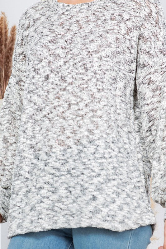 A woman posing in a casual outfit. She's wearing a gray and white patterned sweater and blue jeans. Her left hand is on her head, and her right hand is on her hip. There's a decorative plant with fluffy, tan plumes behind her against a white background.