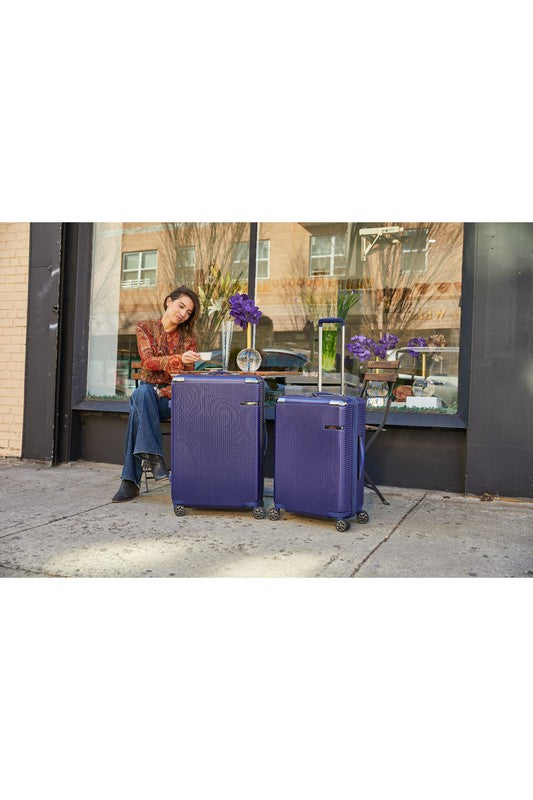 Two purple suitcases with extended handles sit on an outdoor table beside a person, alongside a vase of flowers and a small clock, in front of a reflective storefront window.