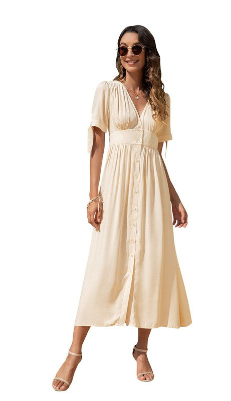 A woman in a cream-colored maxi dress with short sleeves and a V-neckline, wearing sunglasses and strappy heels, standing against a white background.