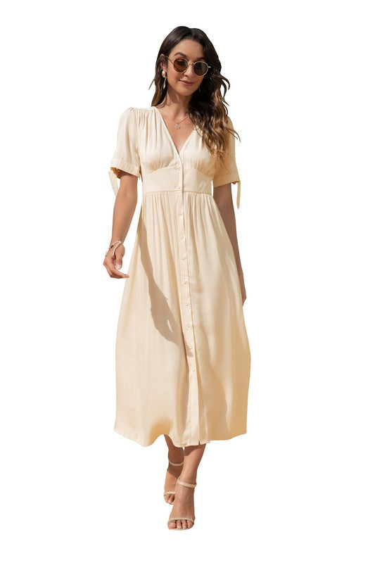 A woman in a cream-colored maxi dress with short sleeves and a V-neckline, wearing sunglasses and strappy heels, standing against a white background.