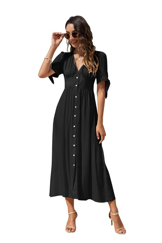 A woman in a black maxi dress with short sleeves and a V-neckline, wearing sunglasses and strappy heels, standing against a white background.