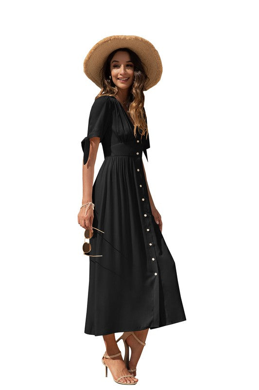 A woman in a black maxi dress with short sleeves and a V-neckline, wearing sunglasses and strappy heels, standing against a white background.