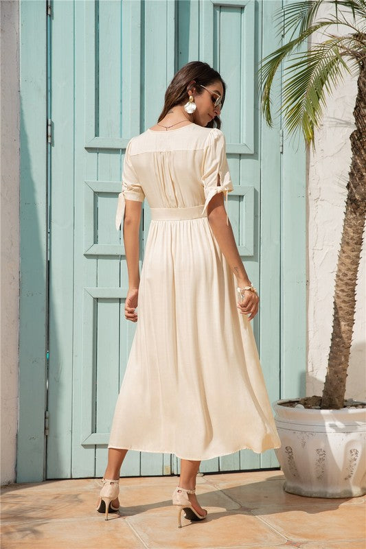 A woman in a cream-colored maxi dress with short sleeves and a V-neckline, wearing sunglasses and strappy heels, standing against a white background.