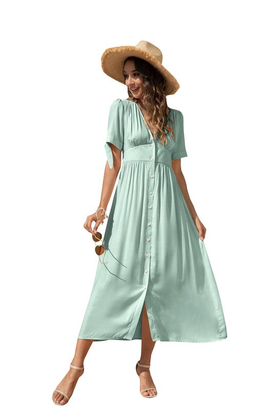 A woman in a cream-colored maxi dress with short sleeves and a V-neckline, wearing sunglasses and strappy heels, standing against a white background.