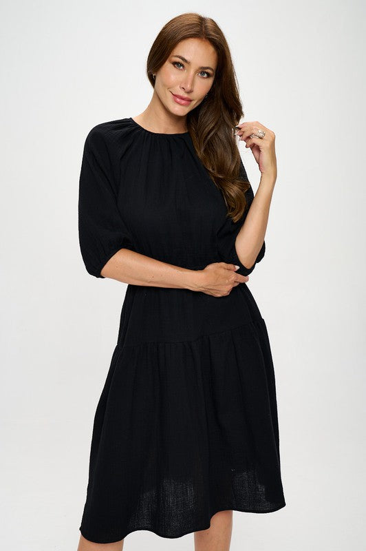 Woman wearing a black midi dress with elbow-length sleeves, gathered waist, and modest flare skirt, styled with black ankle boots.
