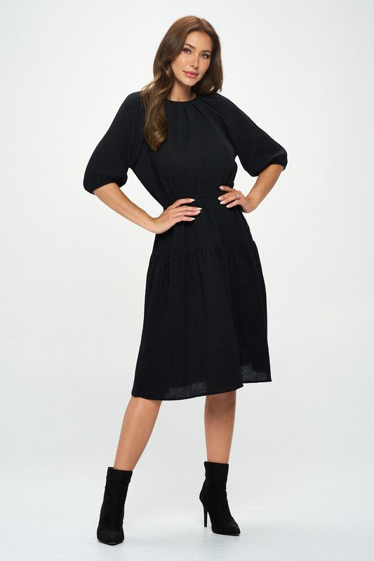 Woman wearing a black midi dress with elbow-length sleeves, gathered waist, and modest flare skirt, styled with black ankle boots.