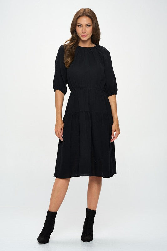 Woman wearing a black midi dress with elbow-length sleeves, gathered waist, and modest flare skirt, styled with black ankle boots.