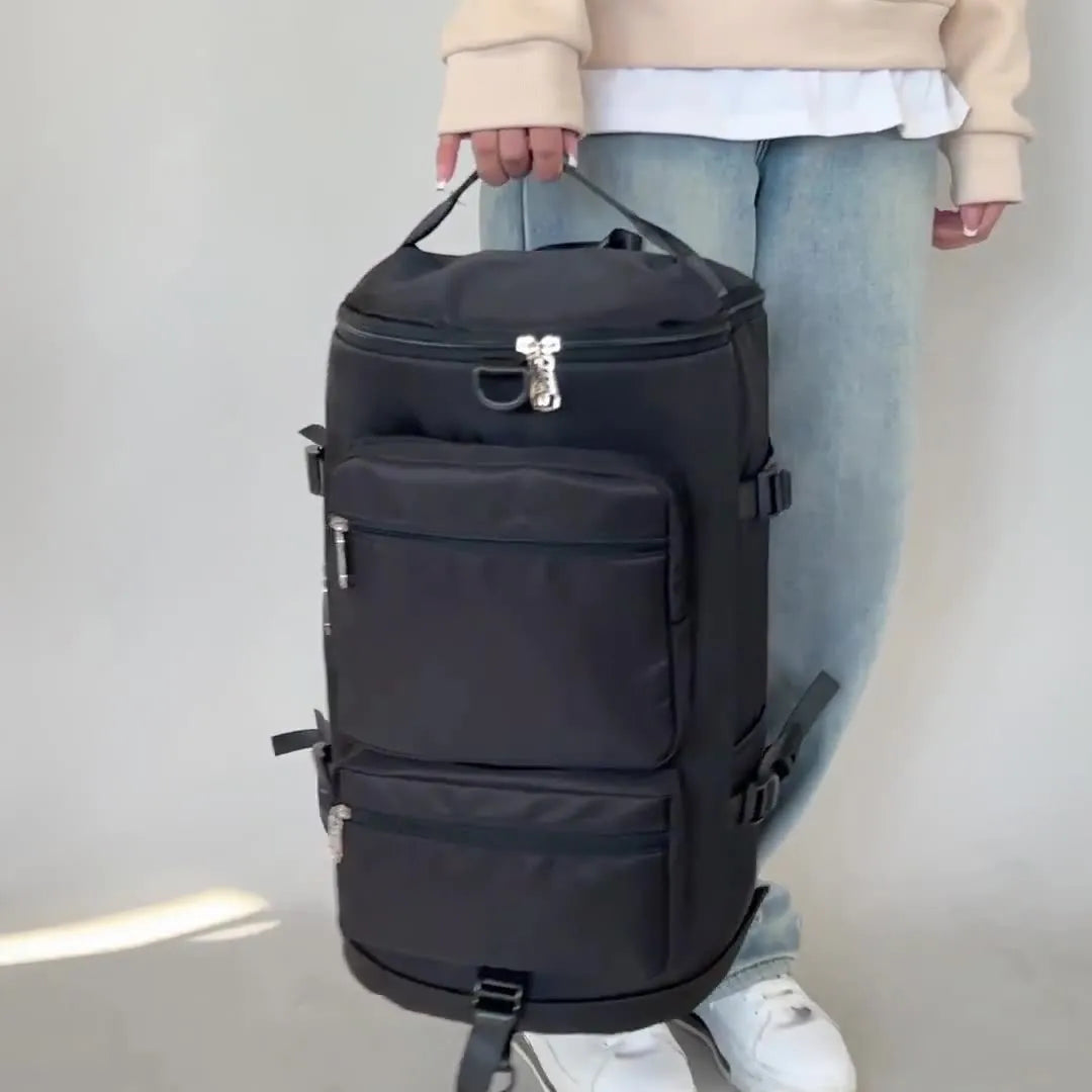Person holding a large black cylindrical backpack with multiple zippered compartments and side straps. Rugged and spacious—ideal for travel or outdoor use.
