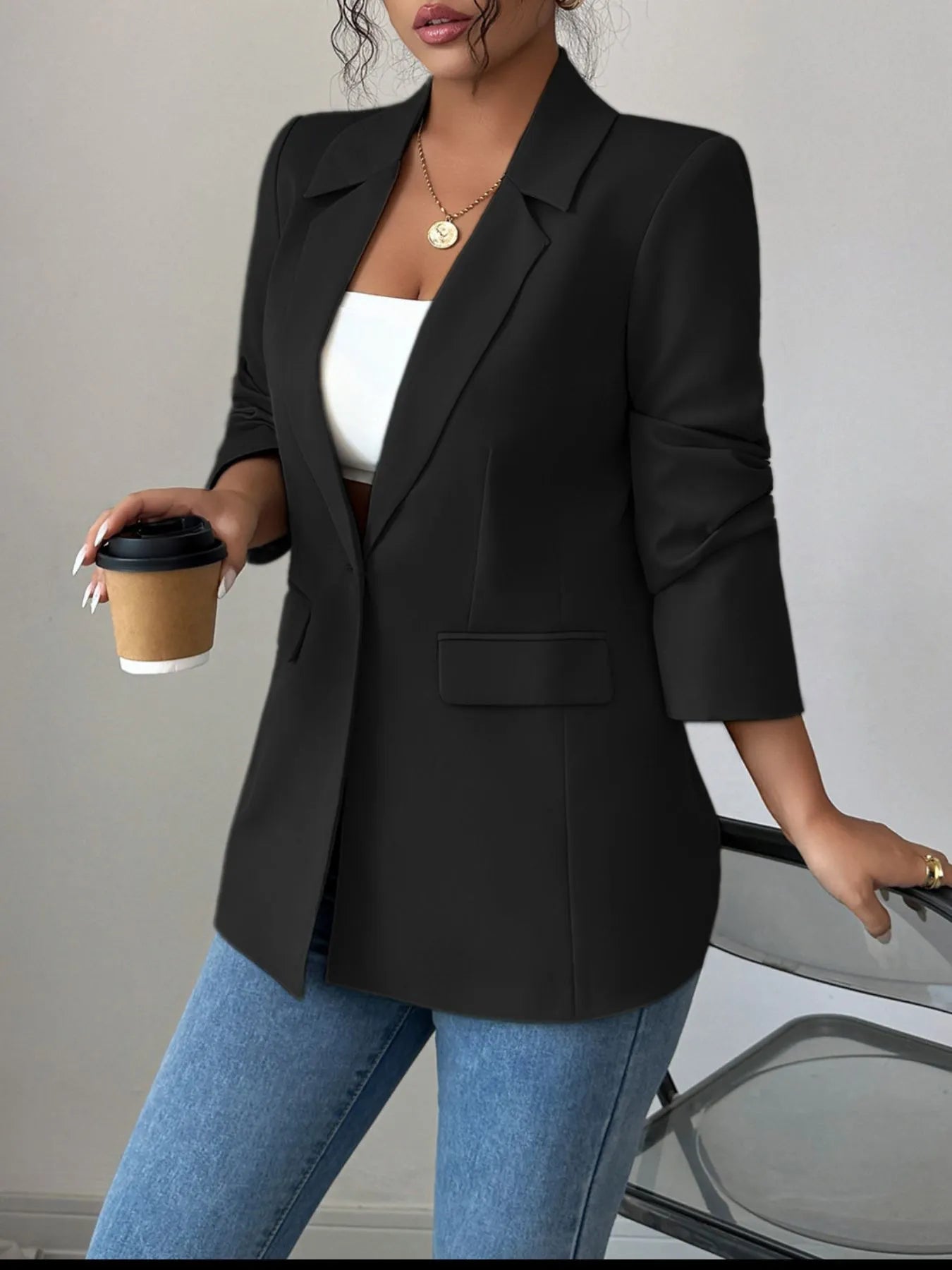 A woman wearing a black blazer, a white crop top, and blue jeans. She's posing with one hand on her hip, showing off some stylish accessories like a gold necklace and rings. The background is a plain gray wall.