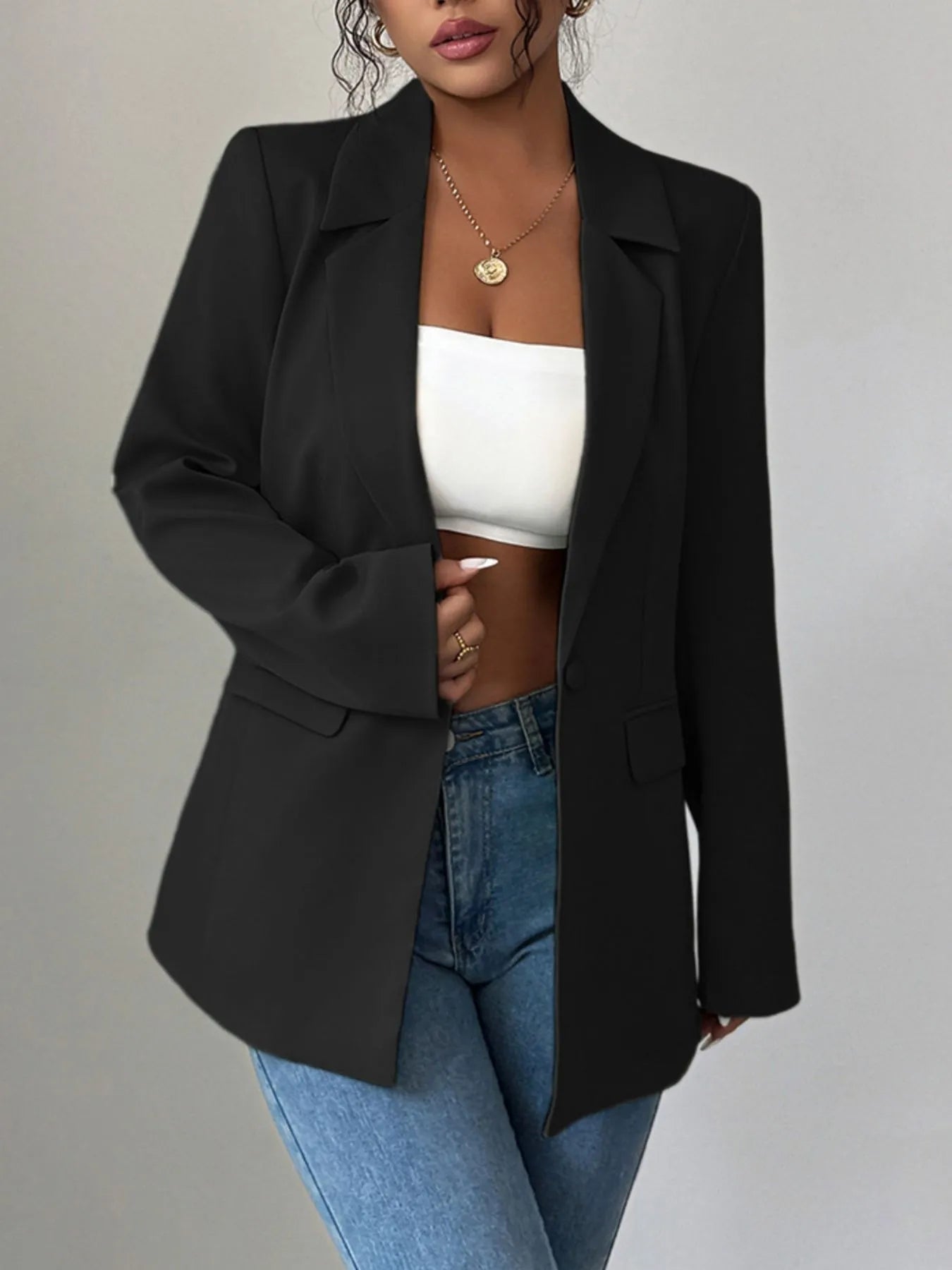 A woman wearing a black blazer, a white crop top, and blue jeans. She's posing with one hand on her hip, showing off some stylish accessories like a gold necklace and rings. The background is a plain gray wall.