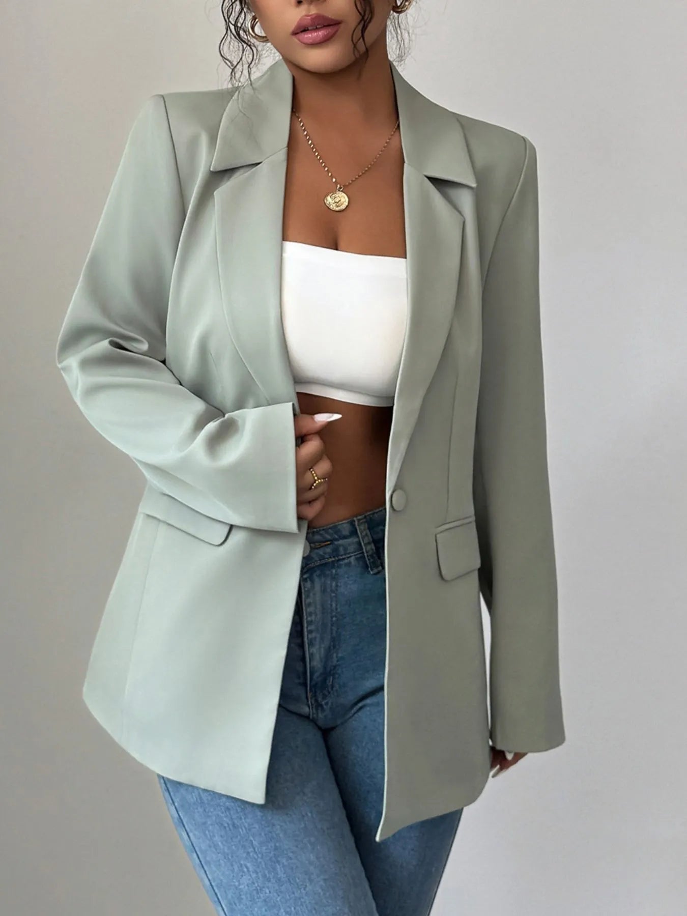 A woman wearing a light green blazer, a white crop top, and blue jeans. She's posing with one hand near her waist and has curly hair. There's a gold necklace with a pendant around her neck.