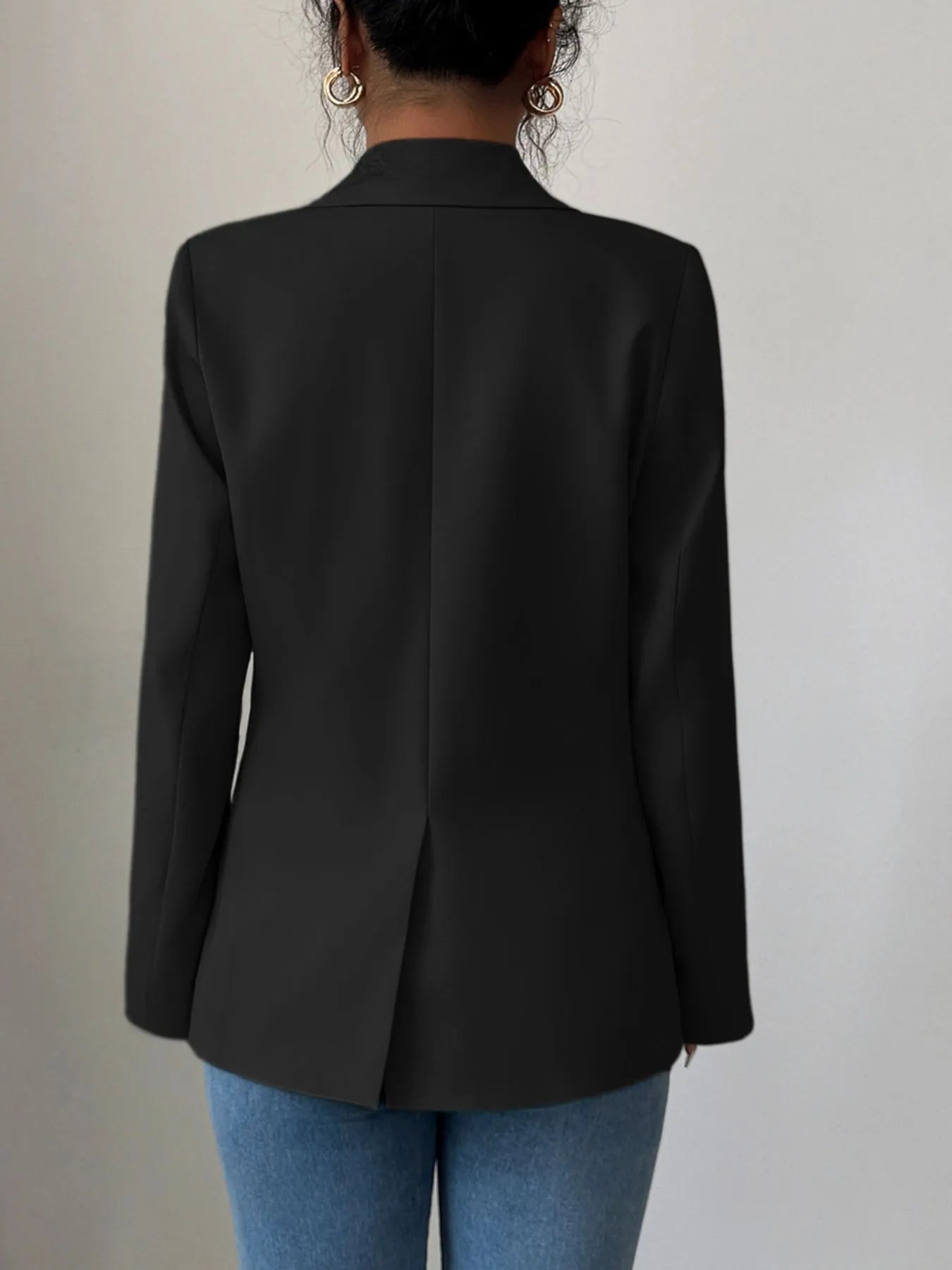 A woman wearing a black blazer, a white crop top, and blue jeans. She's posing with one hand on her hip, showing off some stylish accessories like a gold necklace and rings. The background is a plain gray wall.