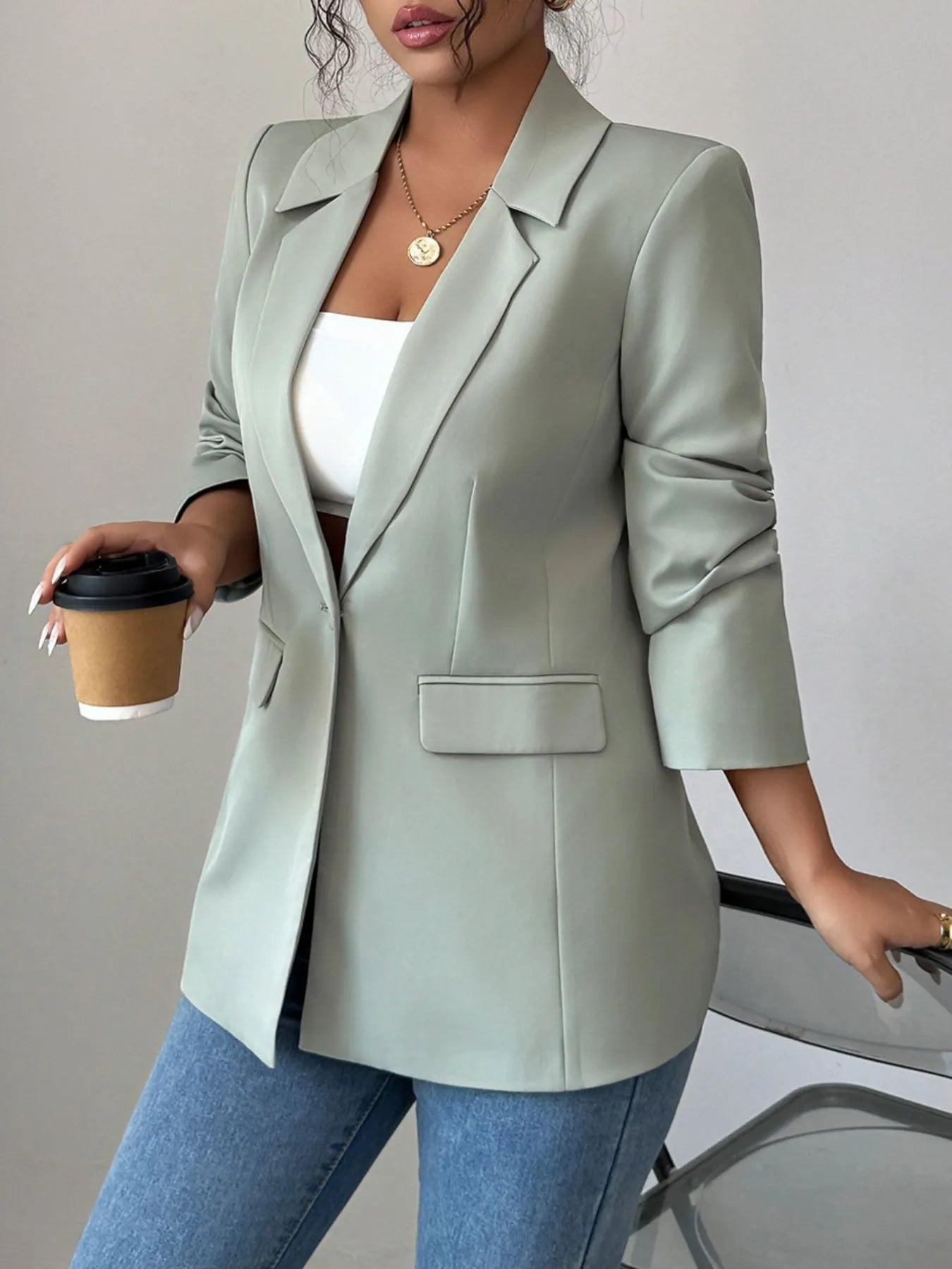 A woman wearing a light green blazer, a white crop top, and blue jeans. She's posing with one hand near her waist and has curly hair. There's a gold necklace with a pendant around her neck.