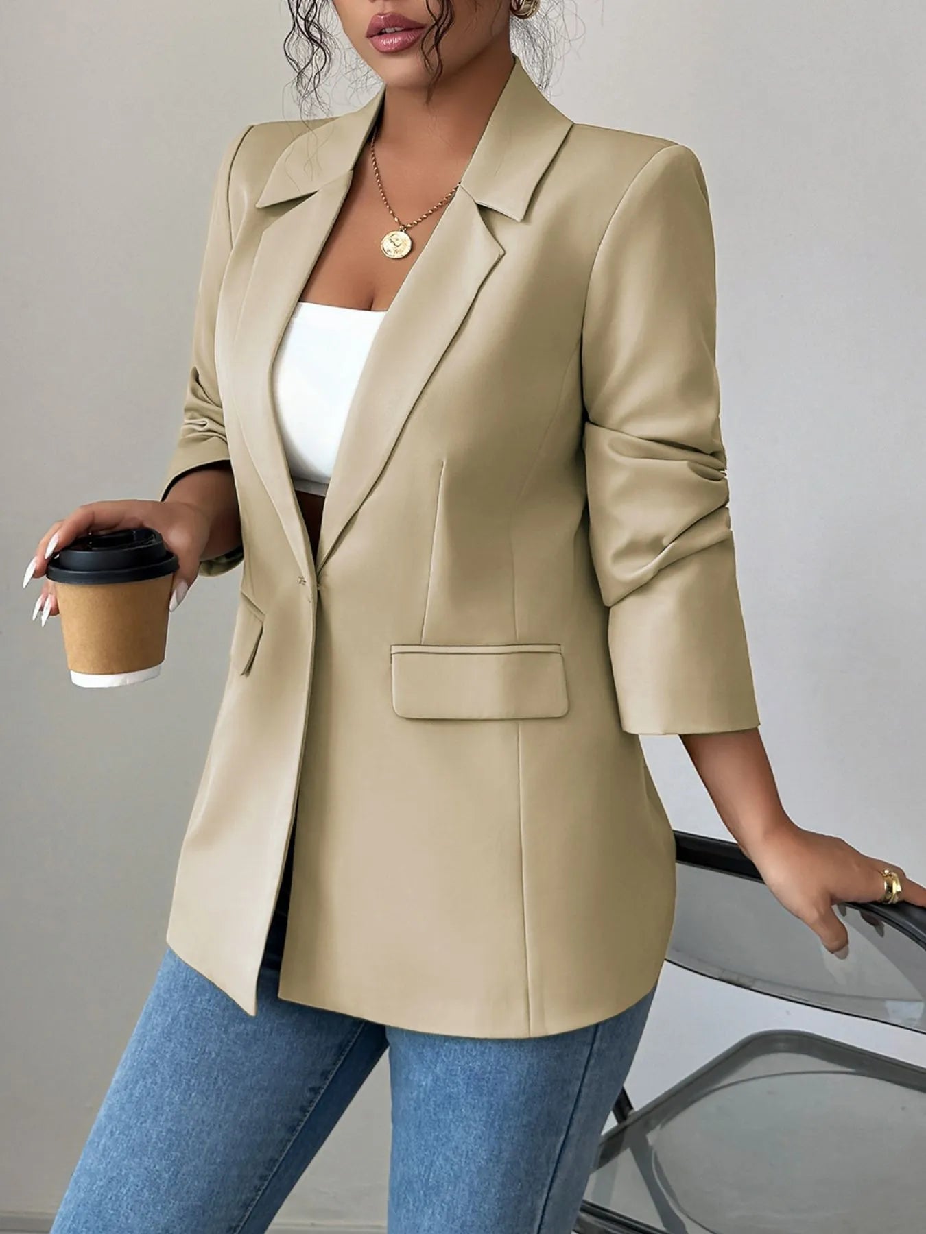 A woman wearing a tan blazer, a white crop top, and blue jeans. She's posing with one hand on her hip, showing off some stylish accessories like a gold necklace and rings. The background is a plain gray wall.