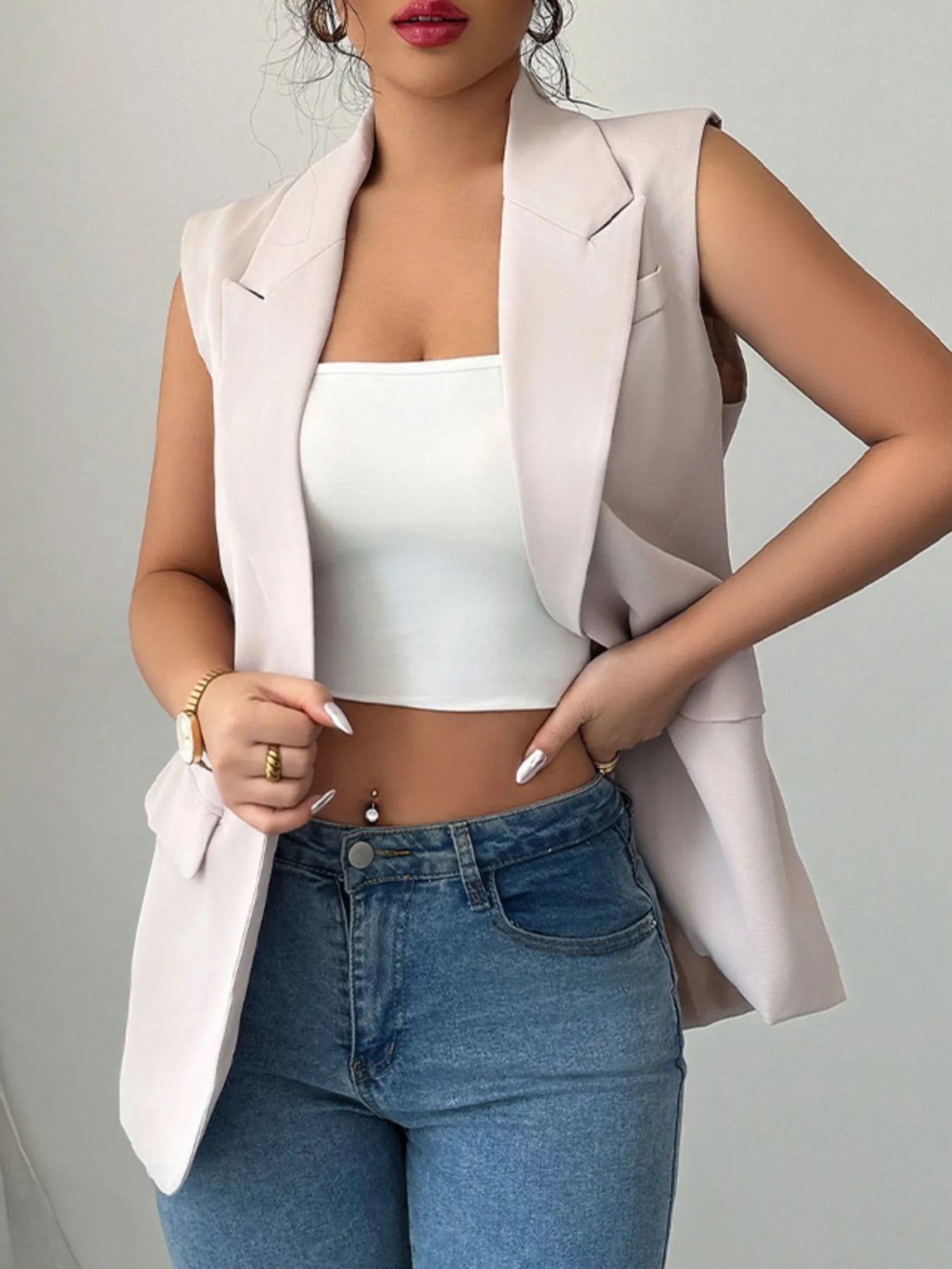 The lady is rocking a sleeveless light pink blazer vest over a white top with blue jeans. She's holding a white purse with a gold chain.