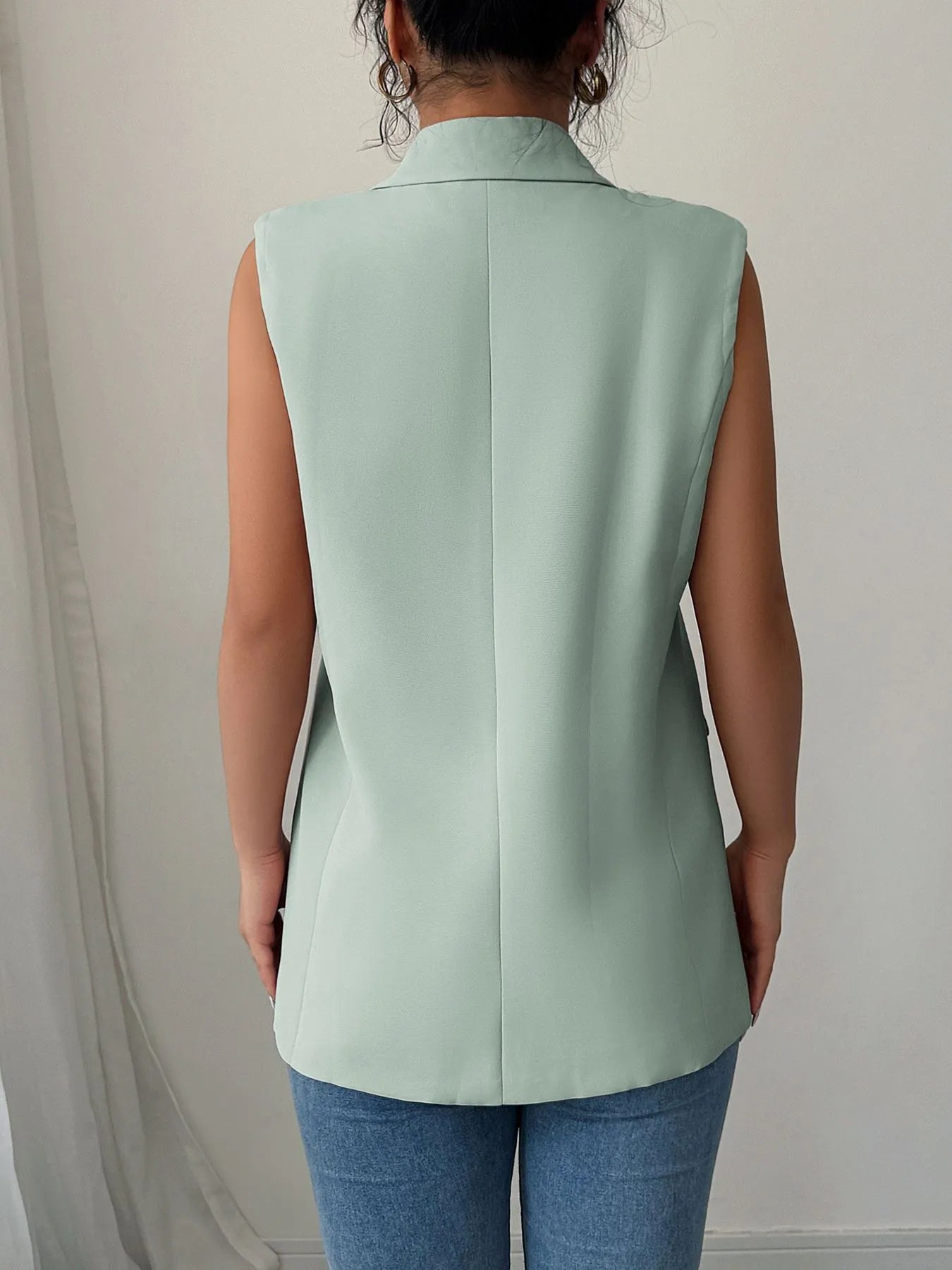 A woman wearing a mint green sleeveless blazer with a white top underneath, paired with blue jeans. She's holding a white purse in one hand and touching her hair with the other.