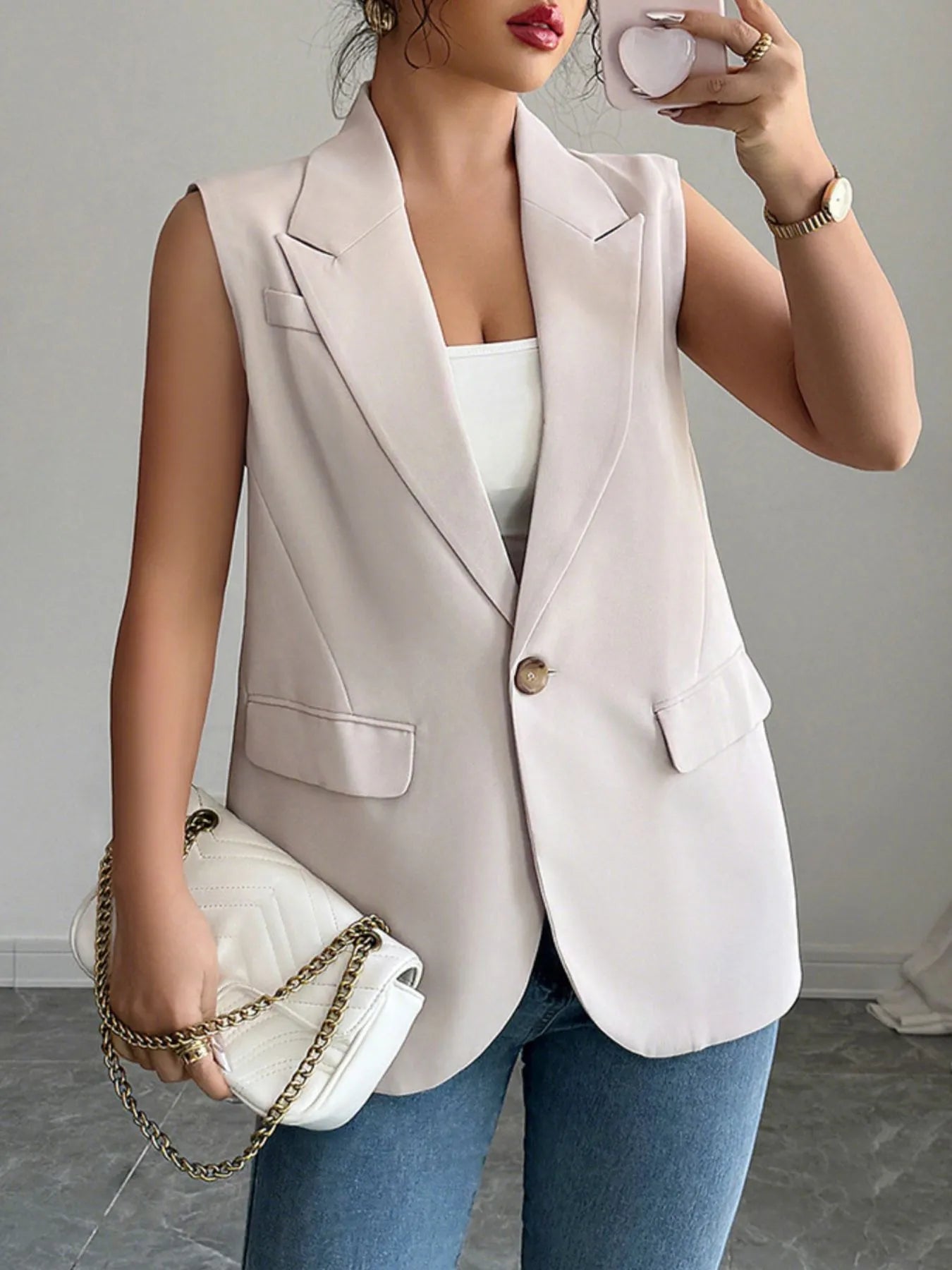 The lady is rocking a sleeveless light pink blazer vest over a white top with blue jeans. She's holding a white purse with a gold chain.