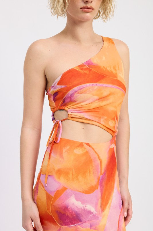 A woman wearing a vibrant one-shoulder maxi dress with an abstract orange, pink, and yellow print. The dress features a cutout at the waist and a slim, straight silhouette, paired with cream knee-high boots and sunglasses