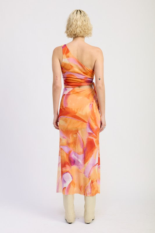 A lady posing on a one-shoulder maxi dress with an abstract orange, pink, and yellow print.