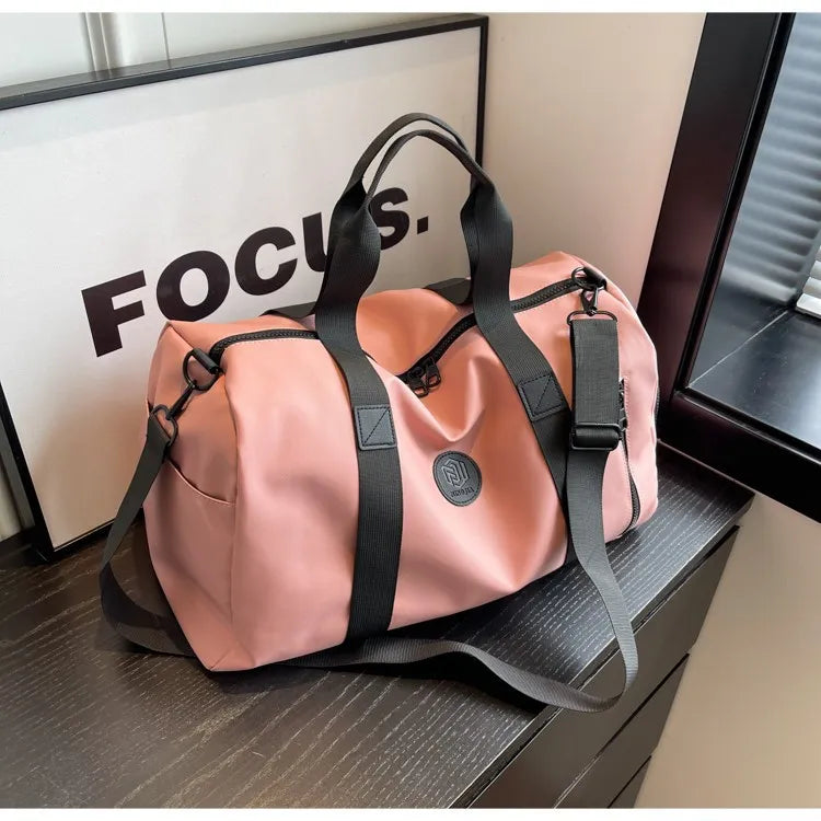 Pink duffel bag with black straps and a circular logo, placed on a dark wooden surface beside a framed "FOCUS" print. Stylish and motivational setup—great for travel or gym use.
