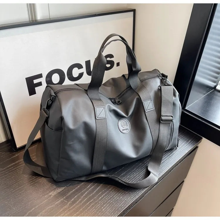 Gray duffel bag with multiple straps and a circular logo, held by a person in a black long-sleeve outfit. Features a zippered side compartment—durable, functional, and ideal for everyday or travel use.