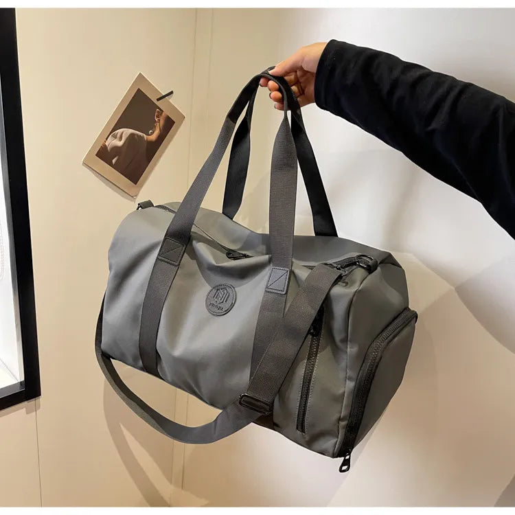 Gray duffel bag with multiple straps and a circular logo, held by a person in a black long-sleeve outfit. Features a zippered side compartment—durable, functional, and ideal for everyday or travel use.